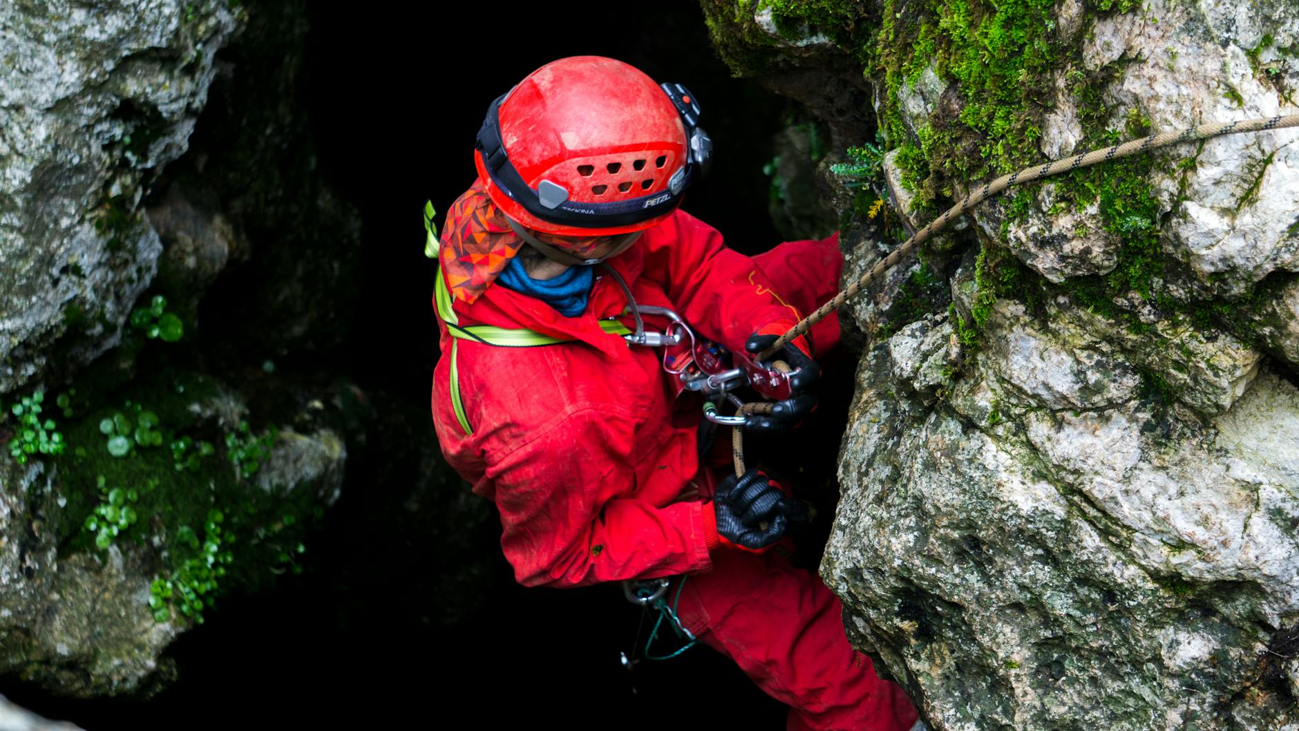 Speleolog w czerwonym kombinezonie pokonuje skalny korytarz jaskini